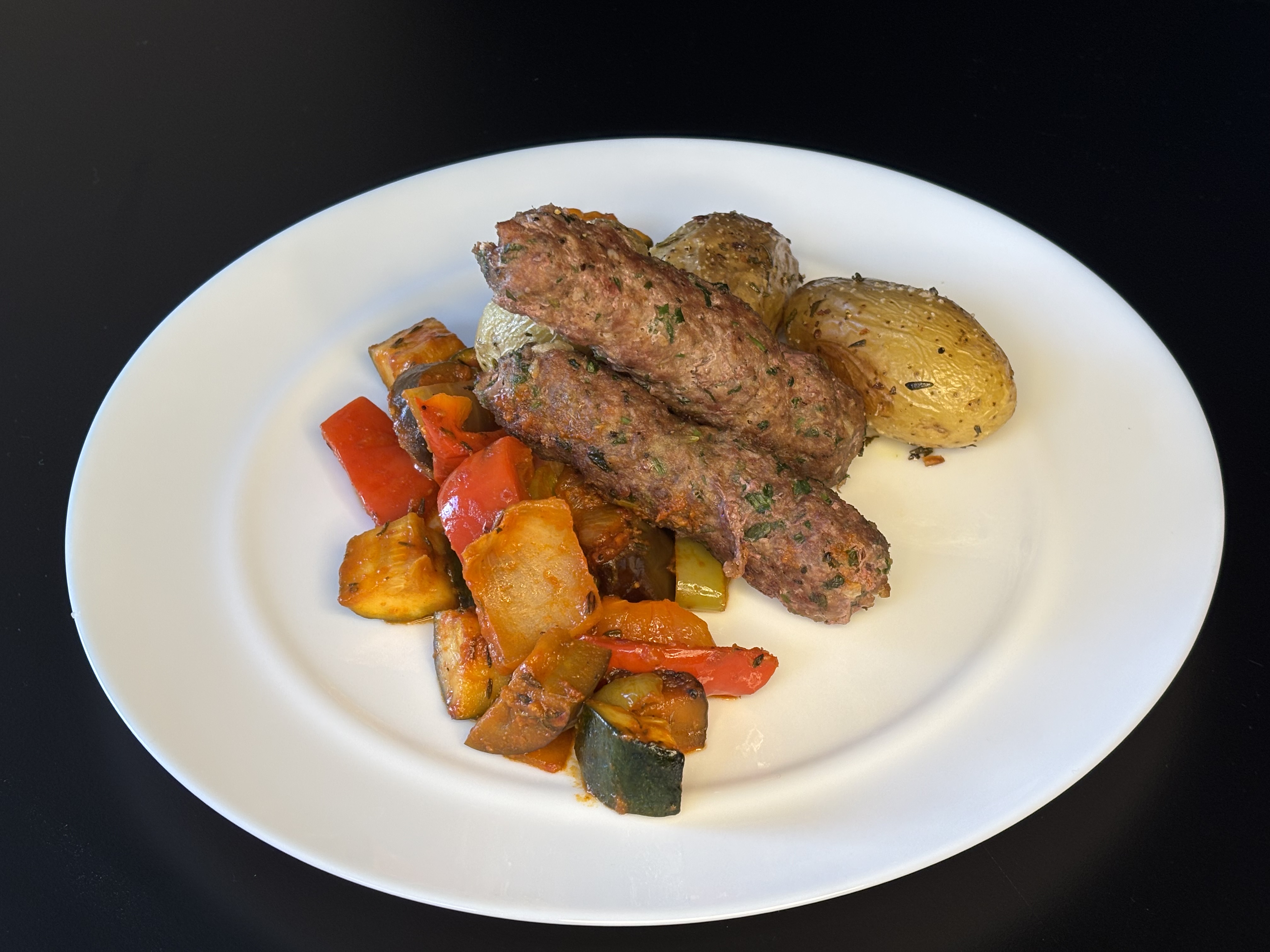 Gegrillte Hackfleischspieße mit gebratenem Gemüse aus Paprika und Zucchini und Kräuterkartoffeln auf weißem Teller.