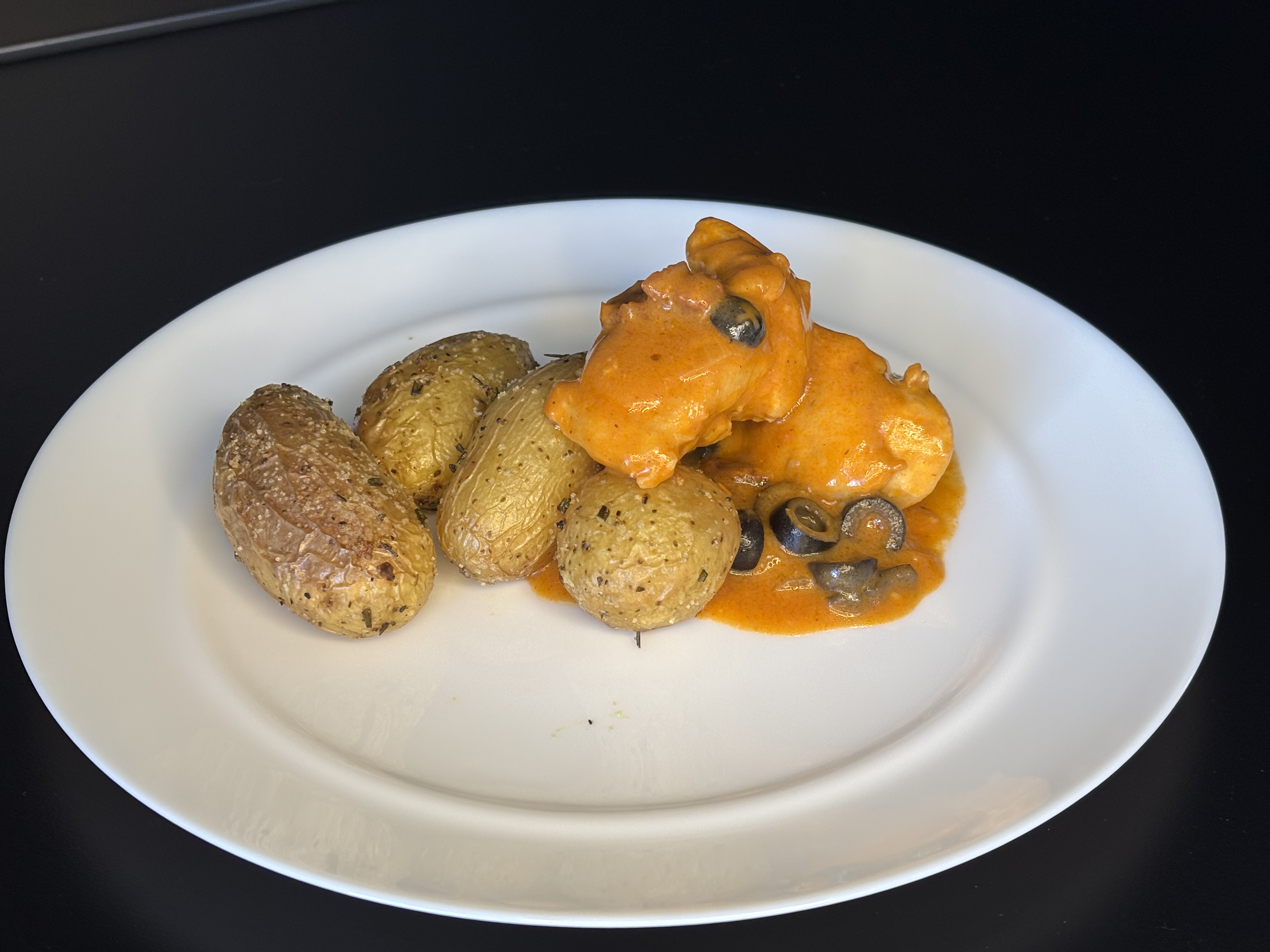 Hähnchenstücke in Tomaten-Oliven-Sauce mit Rosmarin-Drillingskartoffeln auf weißem Teller.