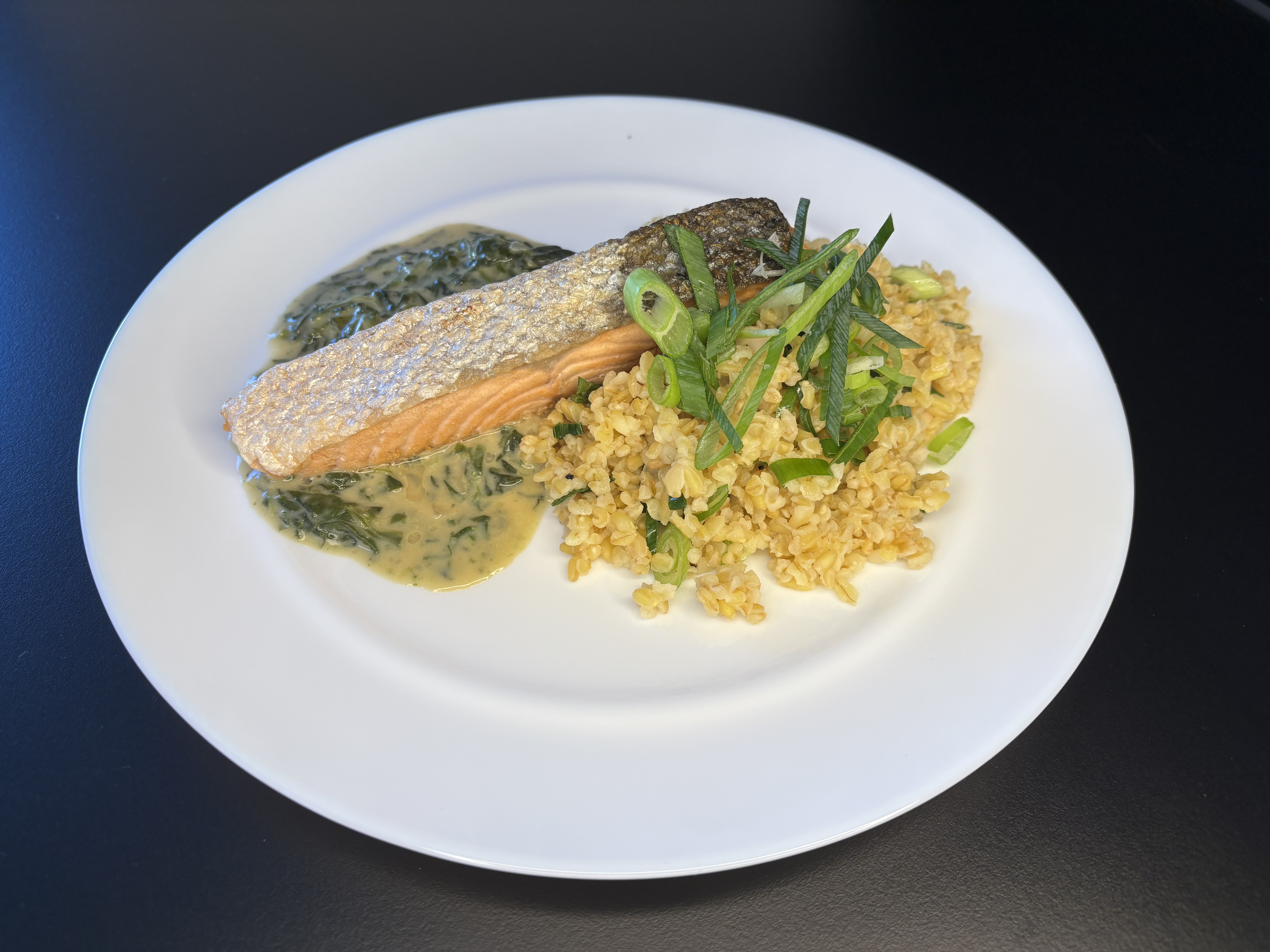 Zart gegarter Fisch auf Spinat mit Bulgur und heller Sauce auf weißem Teller.